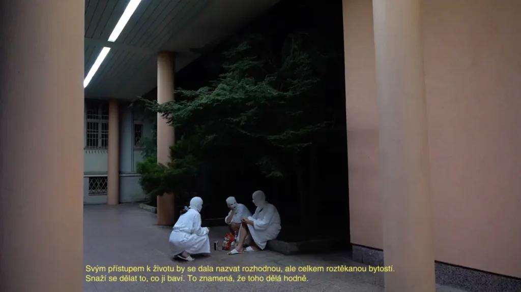 Three figures in white bathrobes, white slippers and white balaclavas are sitting on the pavement in front of the orange painted building at night.