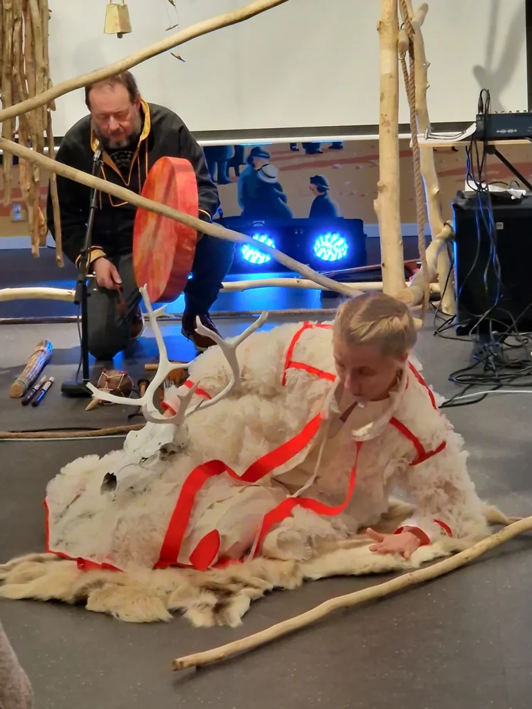 In the image you can see two artists Johan Sara Jr and Katarina Sk&aring;r Lisa. Johan Sara Jr is interacting with drums, while Katarina Sk&aring;r Lisa is holding the white painted reindeer skull.