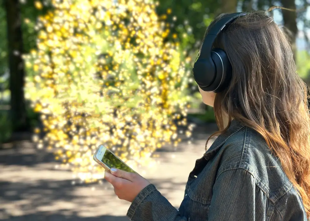 A woman wearing headphones and holding a phone stands outdoors in front of green trees, looking toward a yellow, glittering cloud.