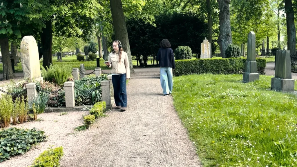 Two young women wearing headphones and holding phones walk among the tombstones in a cemetery during the mixed reality walk Ashes to Ashes.