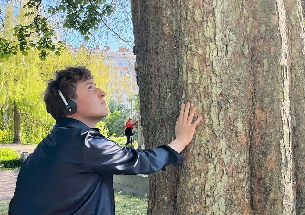 A man wearing headphones places his hand on a tree, listening and looking up during the mixed reality performance Ashes to Ashes.