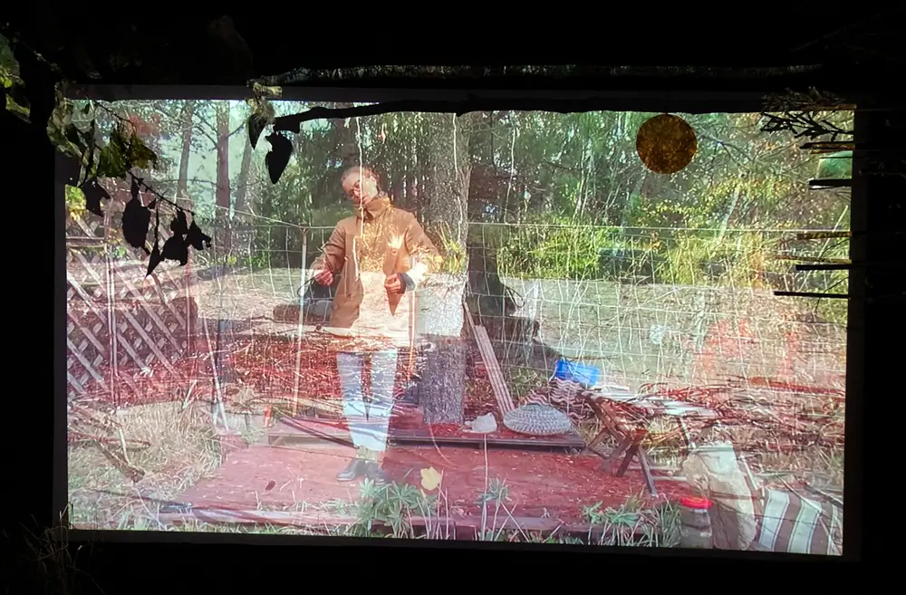 The video work is projected onto the wall in a dark room. Materials such as dried twig, round kombucha-scoby, stick, and reed are installed around the projection. In the projected image, a person taps on a wooden platform in the garden. The picture has two images translucently superimposed; the second image is closer to the legs, which tap on the same platform. Around there is vegetation, such as pine, and some goods, such as a stool and a floor brush.