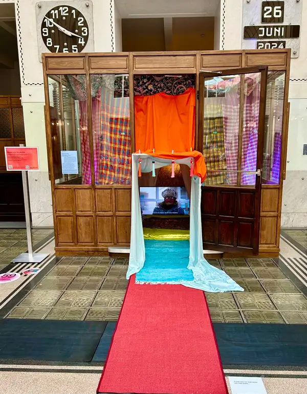 Installation view of &ldquo;Hallabudda Reenactment.&rdquo; A wooden cabin with windows and a central entrance covered in colorful fabrics. A red carpet leads through a blue-framed portal into the interior, where a screen showing a person is visible. The installation is set up inside a large hall.