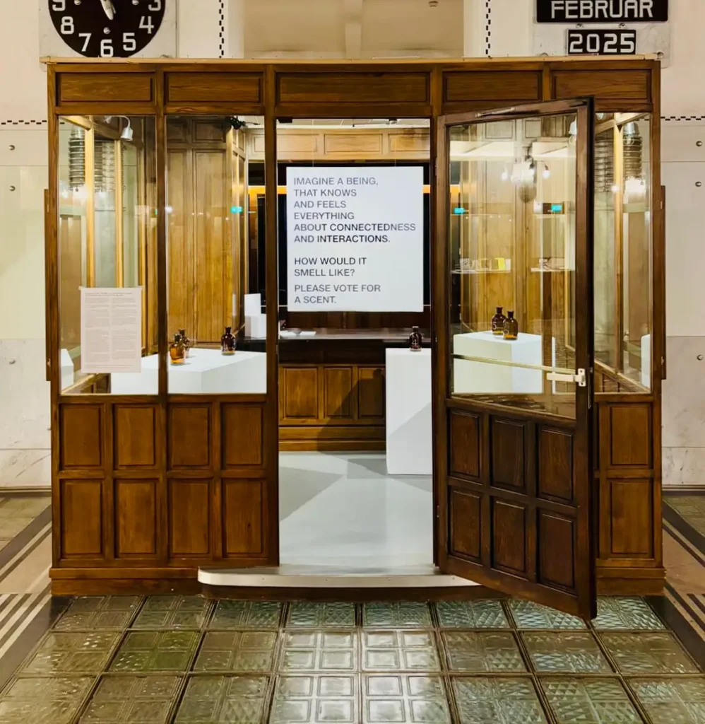Installation view of &ldquo;Conception of SΫS: The Smell of A Mythological Being&rdquo;. A wooden-panelled cabin with windows and an open door reveals several white pedestals bearing in total six brown glass bottles. Behind them hangs a large white poster with black text. It is reading &ldquo;Imagine a being that knows and feels everything about connectedness and interactions. How would it smell like? Please vote for a scent.&rdquo;