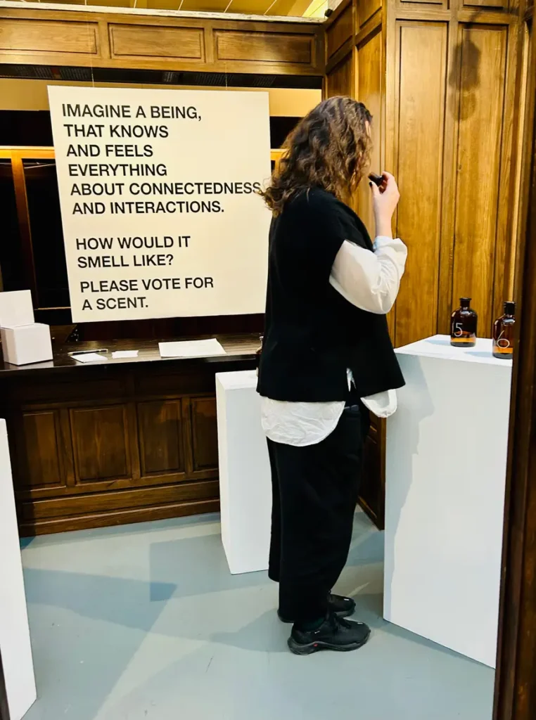 Installation view of &ldquo;Conception of SΫS: The Smell of A Mythological Being.&rdquo; A woman stands inside a wooden-panelled cabin, holding the stopper of a bottle close to her nose. In front of her is a white pedestal with two brown glass bottles. Behind her hangs a poster reading &ldquo;Imagine a being that knows and feels everything about connectedness and interactions. How would it smell like? Please vote for a scent.&rdquo; To the side, a white voting box with paper slips and a pencil is visible.