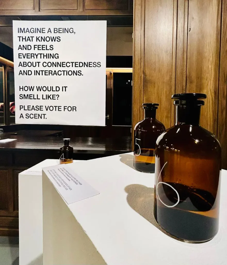 Installation view of &ldquo;Conception of SΫS: The Smell of A Mythological Being&rdquo;. Two brown glass bottles marked with white numbers 5 and 6 stand on a white pedestal. Further pedestals with bottles and a poster printed with black text stating &ldquo;Imagine a being that knows and feels everything about connectedness and interactions. How would it smell like? Please vote for a scent.&rdquo; are visible in the background.