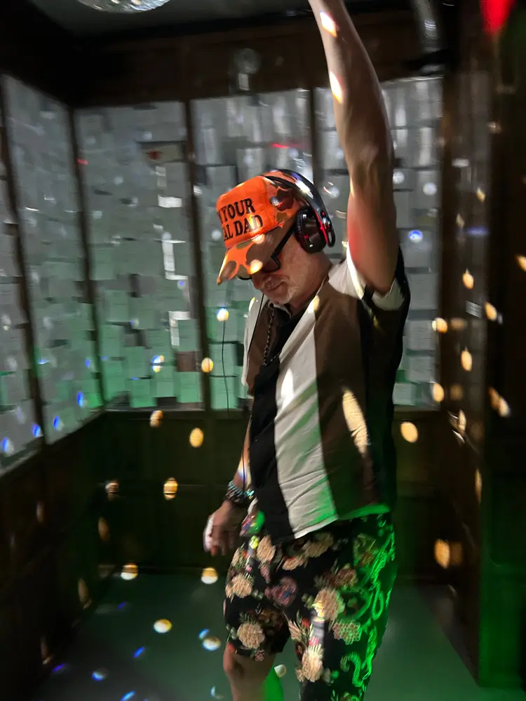 Installation view of &ldquo;SΫS Dance&rdquo;. Interior view with a person wearing headphones dancing. The walls are covered with white paper slips (voting sheets) and illuminated by coloured light.