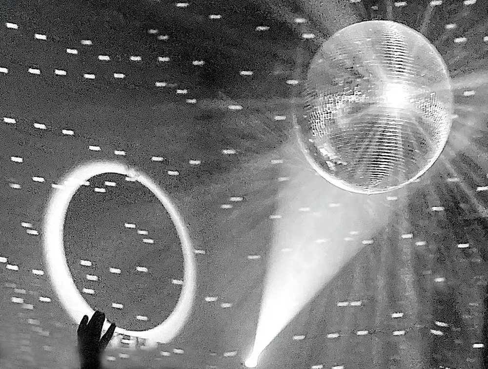Black-and-white image photograph of the installation &ldquo;SΫS Dance&rdquo;. A disco ball reflects bright dots of light onto a dark surface. The outline of a hand appears beside the illuminated area.