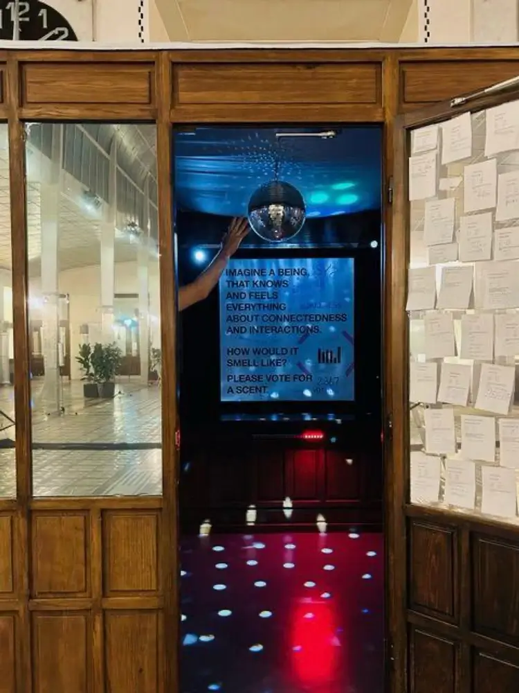 Installation view of &ldquo;SΫS Dance&rdquo;. A wooden-panelled cabin with an open door covered on the inside with numerous white sheets of paper. Inside, a disco ball and a poster with text are visible, while an arm reaches into the frame. The space is lit by blue, red, and white lights.