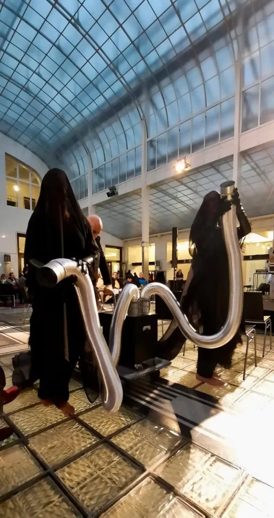 Photograph of the performance &ldquo;Drawing Trees with Scent.&rdquo; Two figures fully covered in black semi-transparent veils stand inside a glass hall. They hold metallic, movable tubes connected to a black device that is pushed between them by a third person in a black suit.