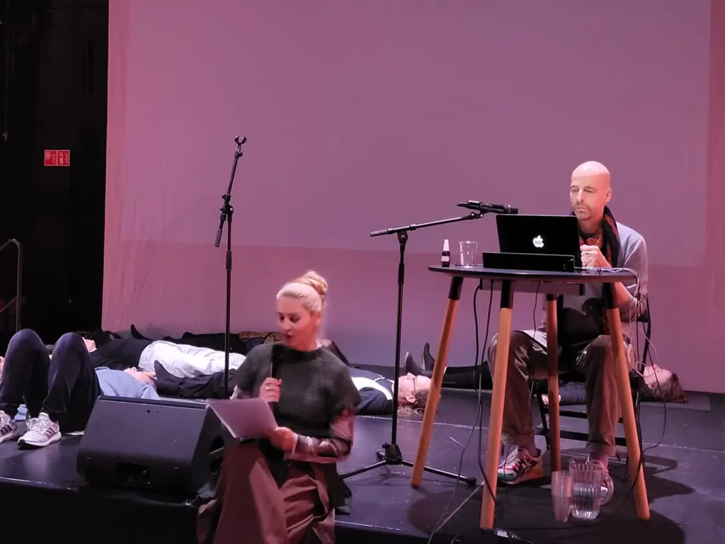Photograph of the multisensory lecture performance &ldquo;Nature After Nature &ndash; Multisensory Medley For Systems Awareness&rdquo; based on Multisensory Imagination Method (MIM) at CARPA9. A woman sits on stage, holding a microphone in one hand and a sheet of paper in the other. Behind her, a man sits at a table with a laptop, facing a microphone on a stand. Next and behind them people are laying on the stage with closed eyes.