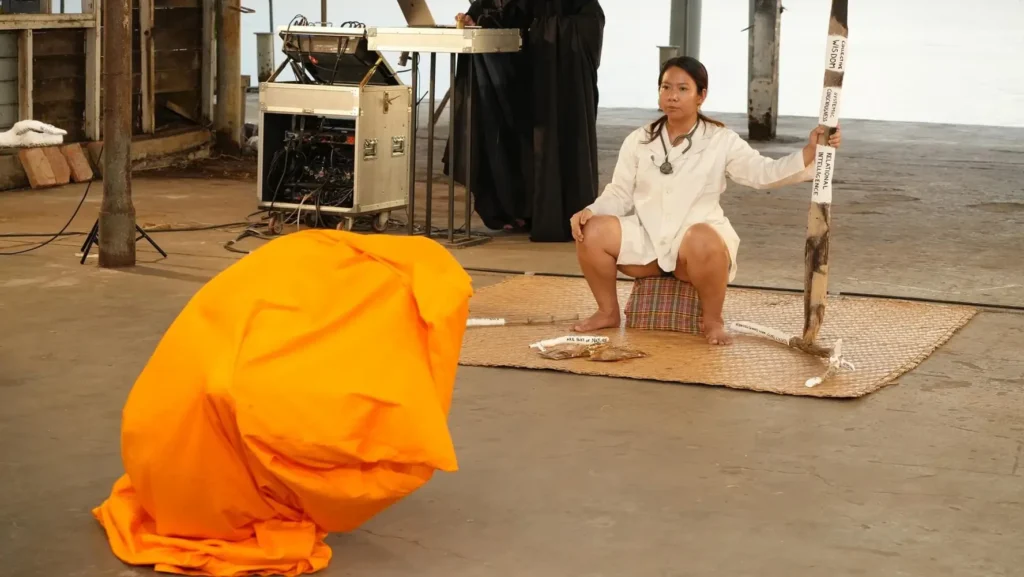 Photograph of the performance &ldquo;Touching Realities&rdquo;. A figure covered with orange fabrics cower on the floor. In the background, a woman in a white lab coat with a stethoscope holds a large branch wrapped with strips of plaster bearing handwritten text. Additional branches lie on a bamboo mat beside her. Behind her, a dark figure stands next to metallic boxes with visible cables.