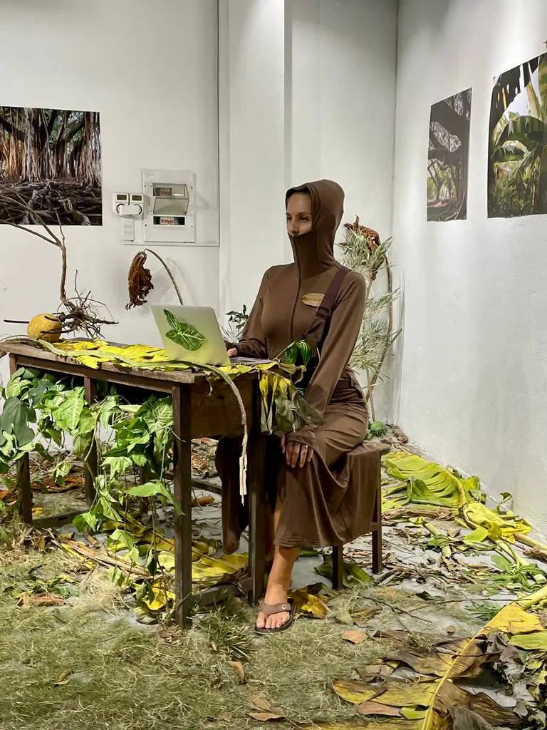 Photograph of the performance &ldquo;Little School of R/Evolution.&rdquo; A person wearing a brown robe and hood sits behind a wooden classroom table overgrown with plants. A laptop stands on the table, while leaves and grass cover the floor. In the background, three images of trees and jungle hang on white walls.