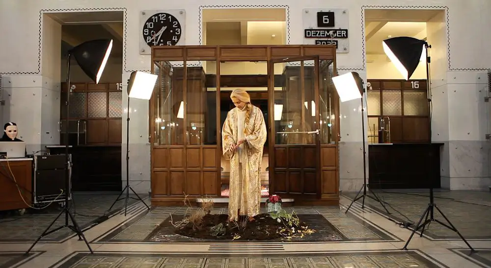 Photograph of the performance &ldquo;Soil Performance.&rdquo; A person wearing a floor-length golden robe stands in a rectangular area filled with soil and growing plants and man-made artifacts. Four spotlights illuminate the scene. In the background, a person wearing a face mask made of plastic sits in front of a laptop. The performance takes place inside a hall with a wooden cabin structure with windows in the background.