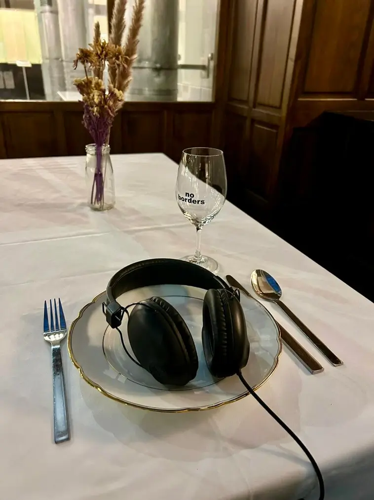Installation view of &ldquo;Expanding System Boundaries&rdquo;. A set table shows a plate with large black headphones placed on top, cutlery beside it, a wine glass labelled &ldquo;no borders&rdquo;, and a glass vase with dried flowers. The background features a wooden-panelled room with a window.&nbsp;