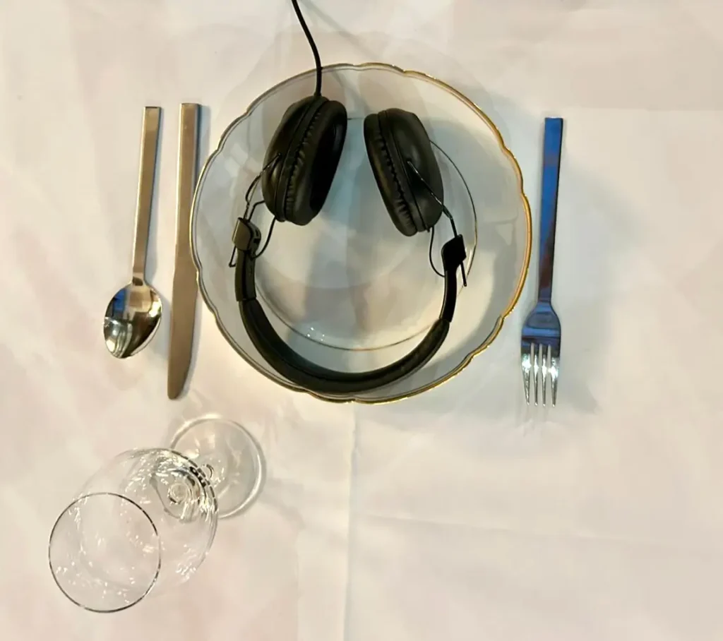 Installation view of &ldquo;Expanding System Boundaries.&rdquo; Overhead view of a set table with a plate holding headphones, cutlery arranged beside it, and an empty wine glass in front of the plate. The headphones resamble a &ldquo;smiley&rdquo; (smiling face emoticon)