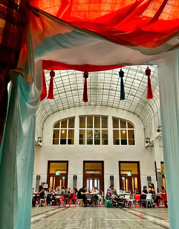 Installation view of &ldquo;Hallabudda Reenactment.&rdquo; View from inside the installation toward the former cashier hall of Otto Wagner Postsparkasse. Through the colorful fabric portal, tables and chairs with people seated are visible.