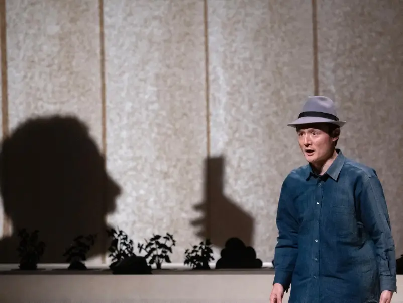 A large shadow of a human side profile projected onto a washi paper background, with a middle-aged man wearing a hat standing on the left side, showing a strong facial expression.