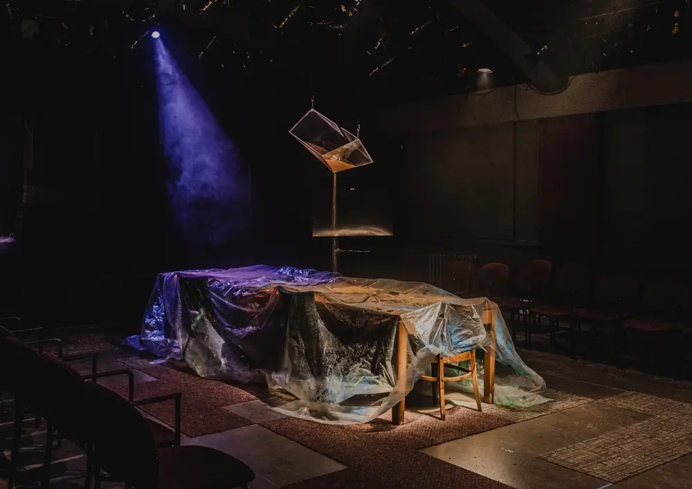 An image of a theatre stage with a long table covered with a plastic sheet. Above the table hangs a glass prism-like object, and sand drips from it.
