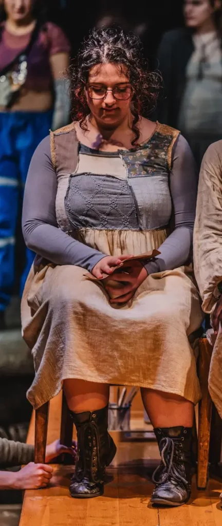 An image of a young woman sitting in a chair, placed at the top of a table. She is wearing a yellow and blue dress.