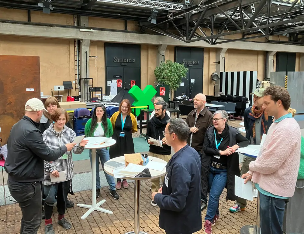 A group of people stands around small tables in the entrance hall at TeaK, listening and smiling as two participants present their workshop results.