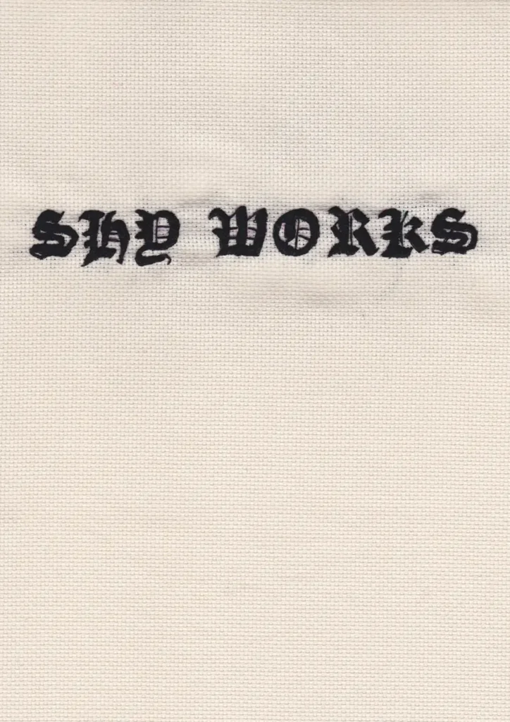 Hand embroidered lettering of the words SHY WORKS in gothic font, on off white canvas.