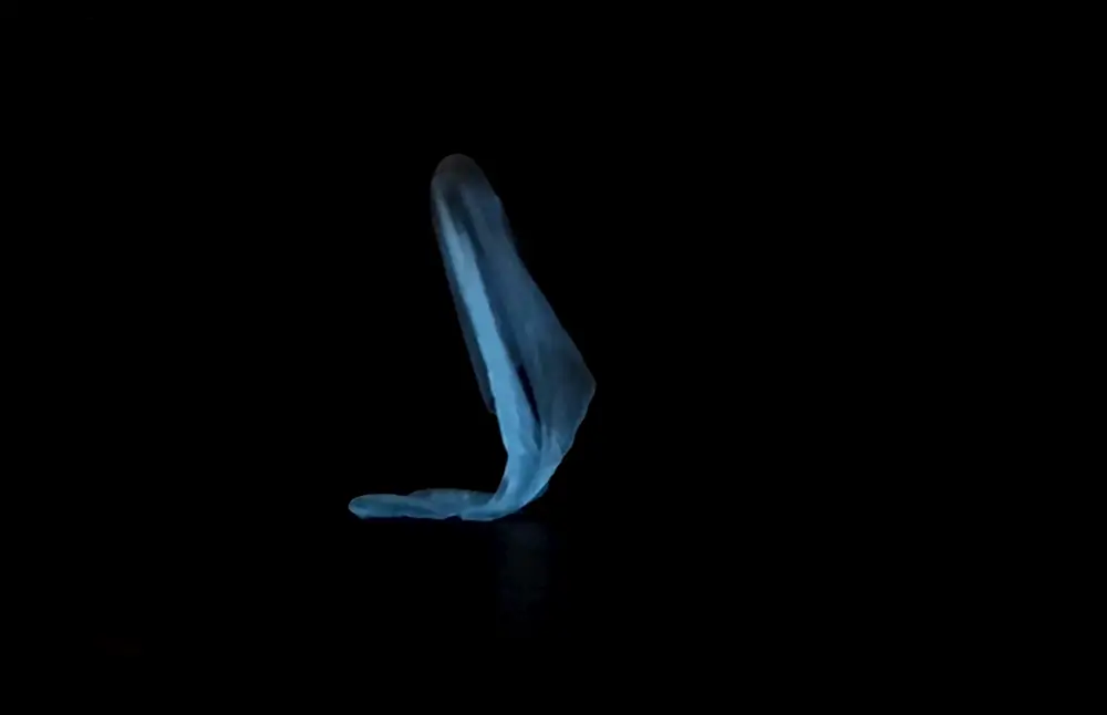 A light blue, semi-transparent fabric form is suspended against a black background. Its shape curves downward toward the floor, with no visible human body or facial features.
