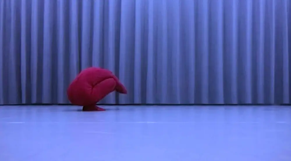 A red, rounded fabric-covered figure rests low to the ground in front of floor-to-ceiling curtains. The shape suggests a compact, folded posture, while the surrounding space is softly lit.