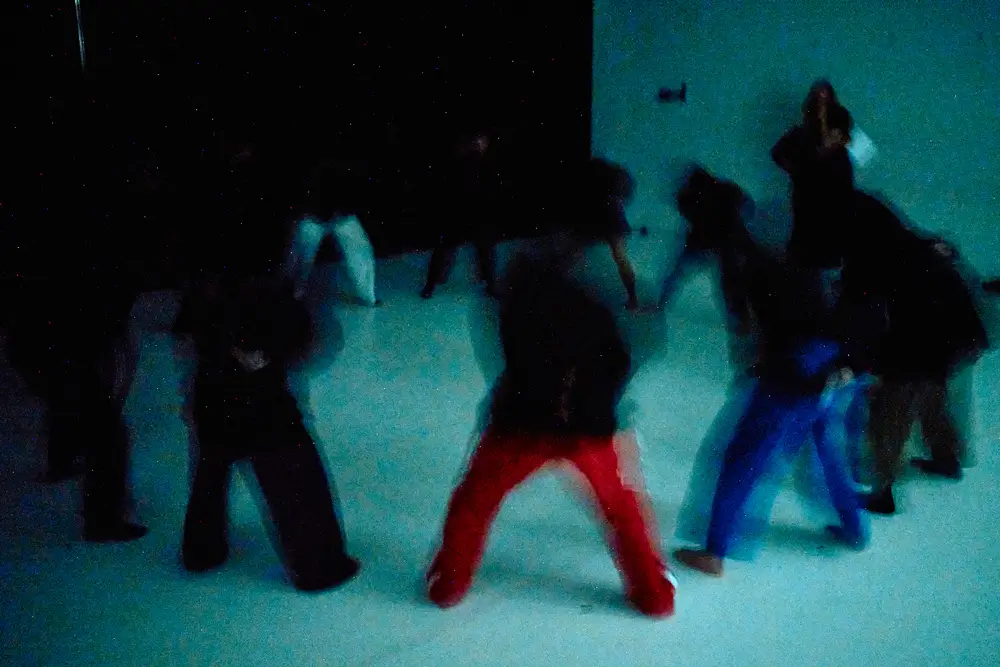 A group of dancers is positioned in a dark studio, wearing black hoodies. Their bodies are slightly blurred in motion, with each person bent forward and feet apart, suggesting synchronized physical engagement.