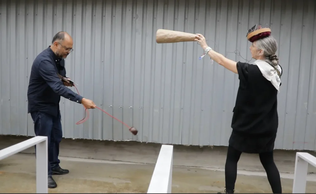 Alejandro Olarte and Marianne Decoser-Taivalkoski improvising with respectively the Feedback Coconut and the Solar Head Tray in front of a grey metallic wall of the yard.