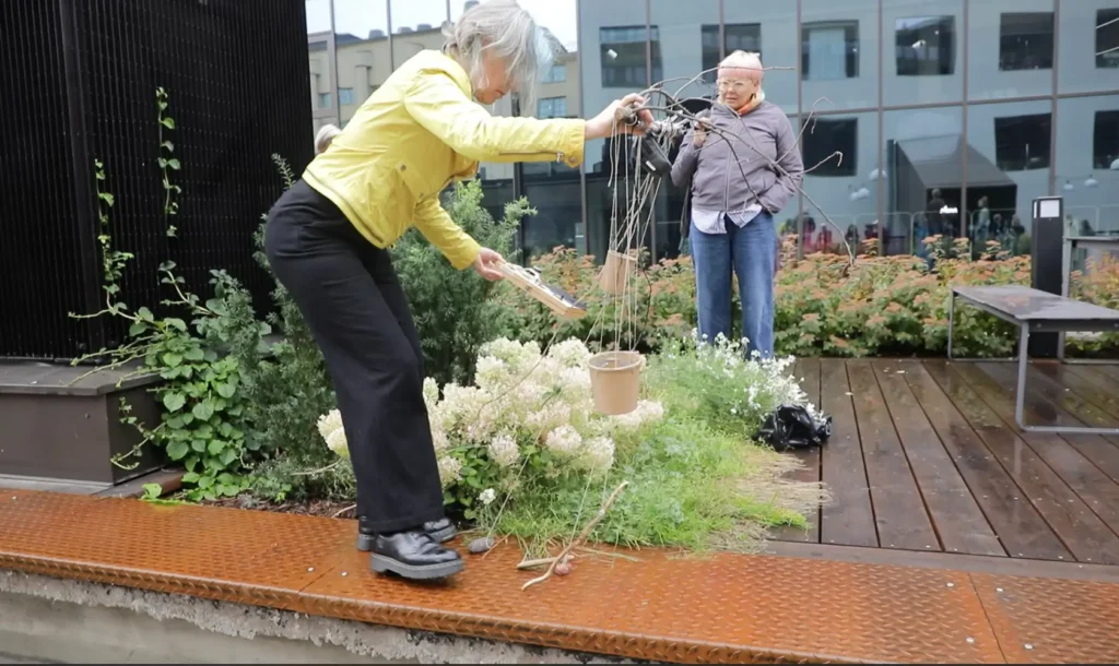 Pascale Weber playing the Cup Harp against a metallic part of the floor of the yard close to a flower bench. An audience member stands right behind.