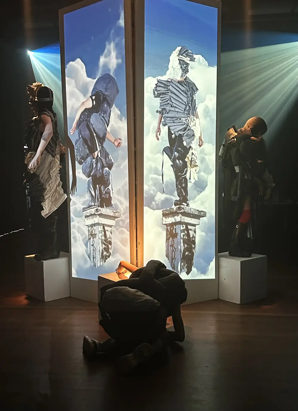 An installation featuring five figures dressed in dark, textured, deconstructed clothing. The two central figures are displayed on tall, vertical screens with a cloudy sky background, standing on digital stone pedestals and seemingly floating in the clouds. Two physical performers stand on white pedestals on either side of the screens, one is crouching in the middle on the floor.