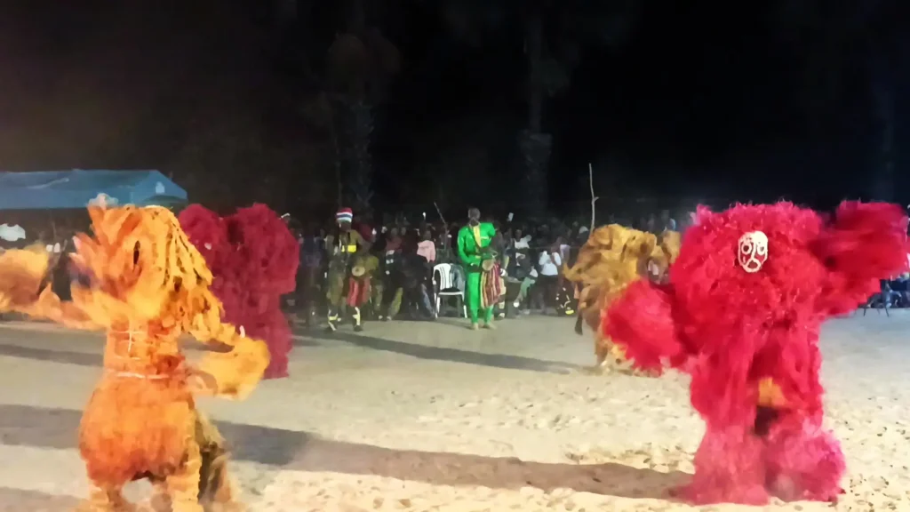 Masquerades performing at night; the one on the front left, the ifangbondi, is made from natural tree bark, and the kankurang masquerade on the front right is bright red and made from synthetic material.