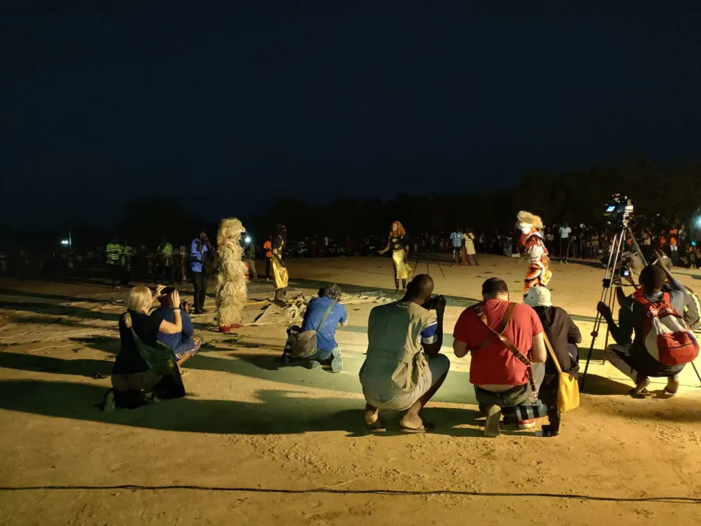 Several photographers and videographers kneel at the edge of an open arena at night, surrounding and capturing a performance featuring the zimba masquerade, with a large audience watching in the background