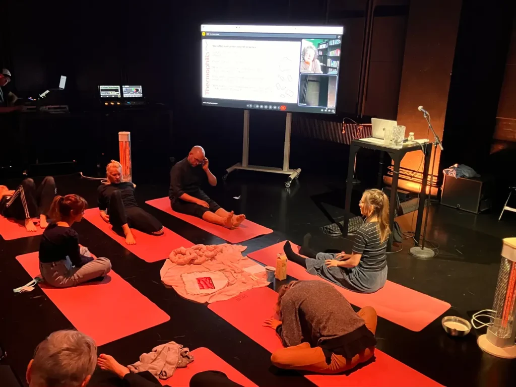 group of people sitting and lying on yoga mats in a dark room, with a large screen behind them, several heaters and bowls