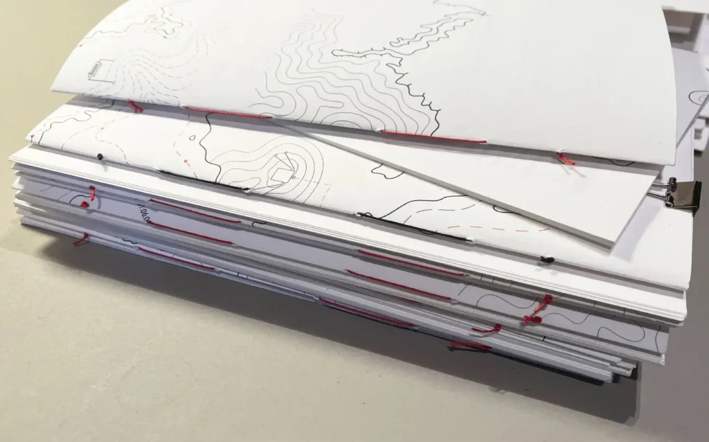 A stack of handmade booklets lies on a table, their covers showing printed contour lines and red thread bindings along the edges.
